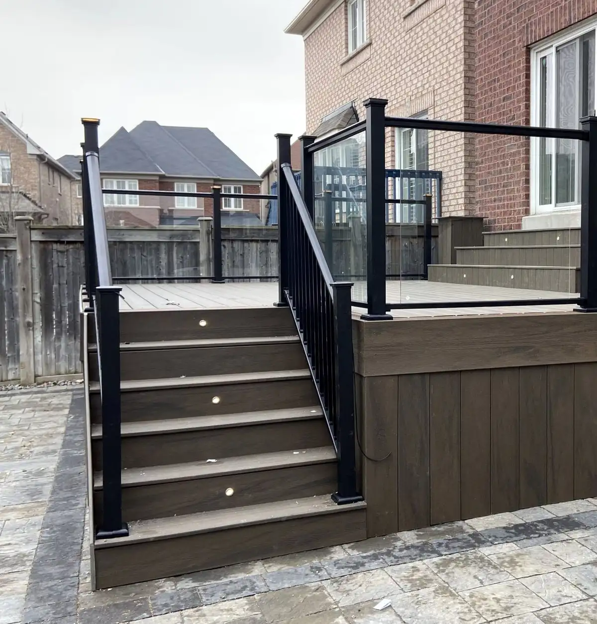 Brown composite stairs with small lights set into the risers and black aluminum railing, rising to a deck with glass railing panels, beside a brick house.