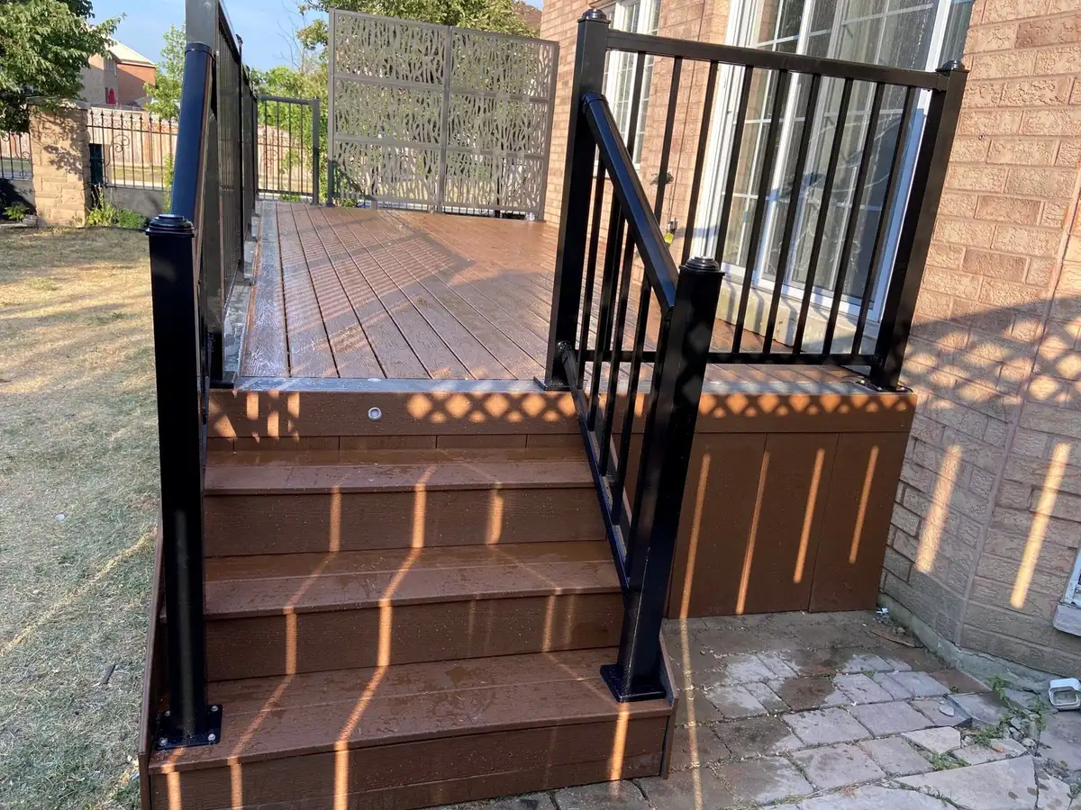 A small brown composite deck with three steps and black aluminum railings beside a brick house, with lattice skirting below.
