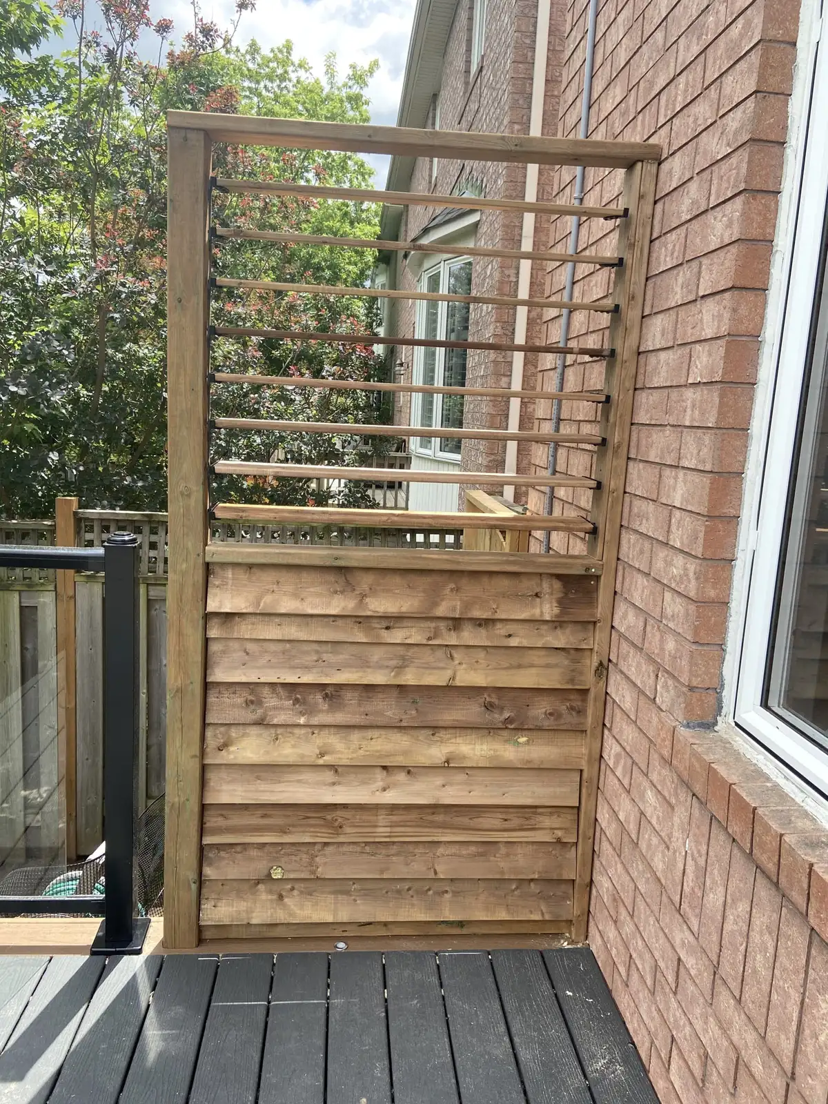 A tall wood privacy wall of horizontal boards with an open slatted top section, standing on a grey deck next to a brick wall and a glass railing.