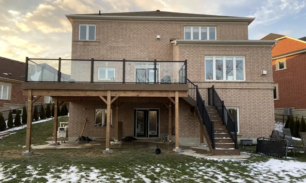 At sunset, a raised deck with glass railing across the back of a large buff-brick house, on timber posts, with a stair with black railings down to the lawn.