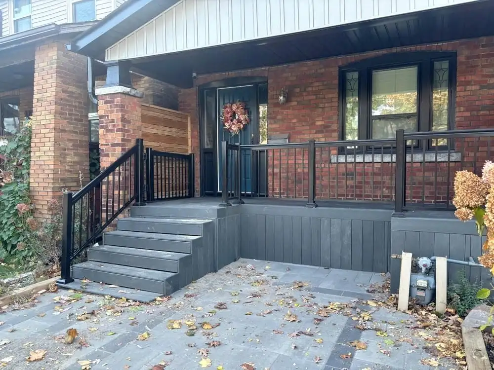 The front porch of a red-brick bungalow rebuilt with charcoal composite steps and skirting and black aluminum railings.