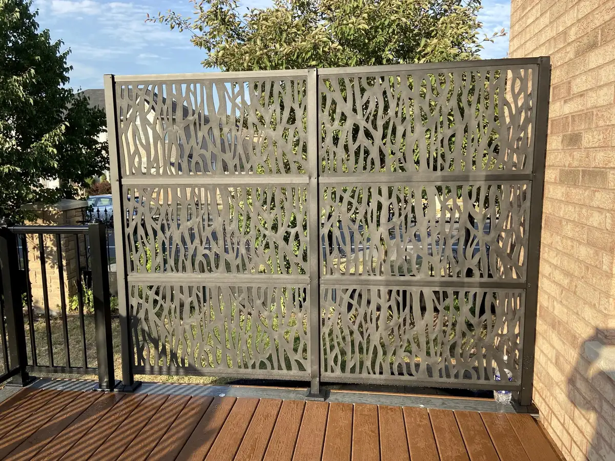Two tall bronze-coloured metal privacy panels with a leaf-like laser-cut pattern, standing on a brown composite deck beside a brick wall.