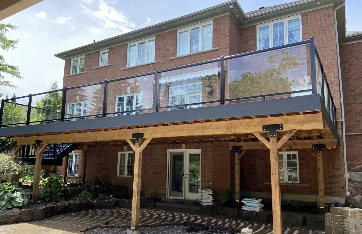 A raised charcoal composite deck along the back of a red-brick house, with glass railing panels between black aluminum posts, carried on timber posts, beams and angled knee braces with black steel brackets.