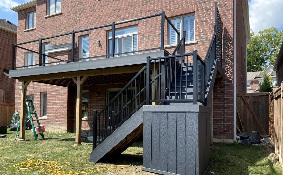 A raised grey composite deck across the back of a brick house, with black aluminum railings, a stair in two flights and a grey composite box at its foot.