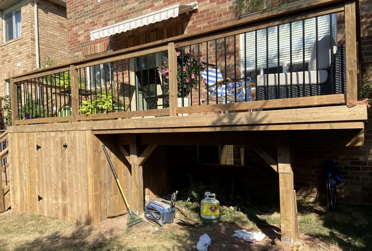 A raised pressure-treated deck with black balusters across the back of a brick house, its underside closed in with vertical wood boards.