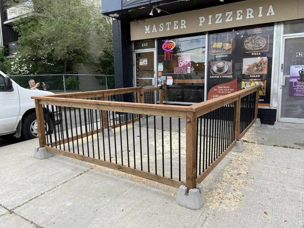 A freestanding patio railing of wood posts and rails with black balusters, enclosing a sidewalk patio in front of a pizzeria.