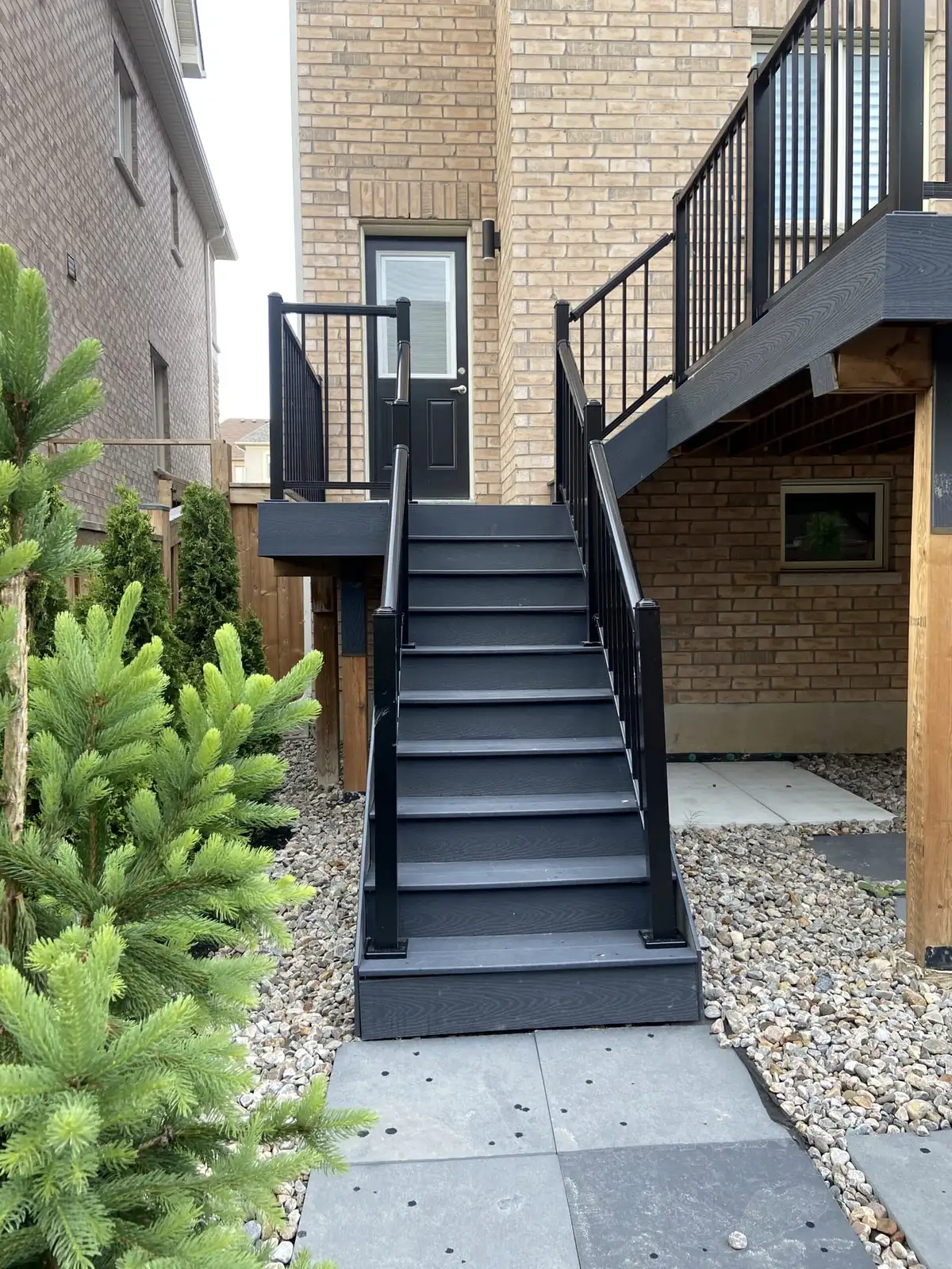 A tall straight stair in charcoal composite with black aluminum railings, climbing along a buff-brick house to a raised deck, with gravel and stone pavers at its foot.