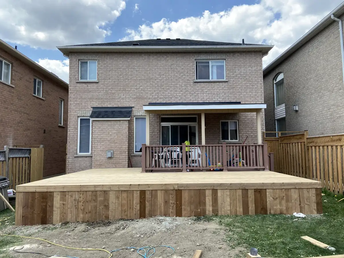 A ground-level pressure-treated platform deck spanning the back of a two-storey brick house, with a short railing at the door.