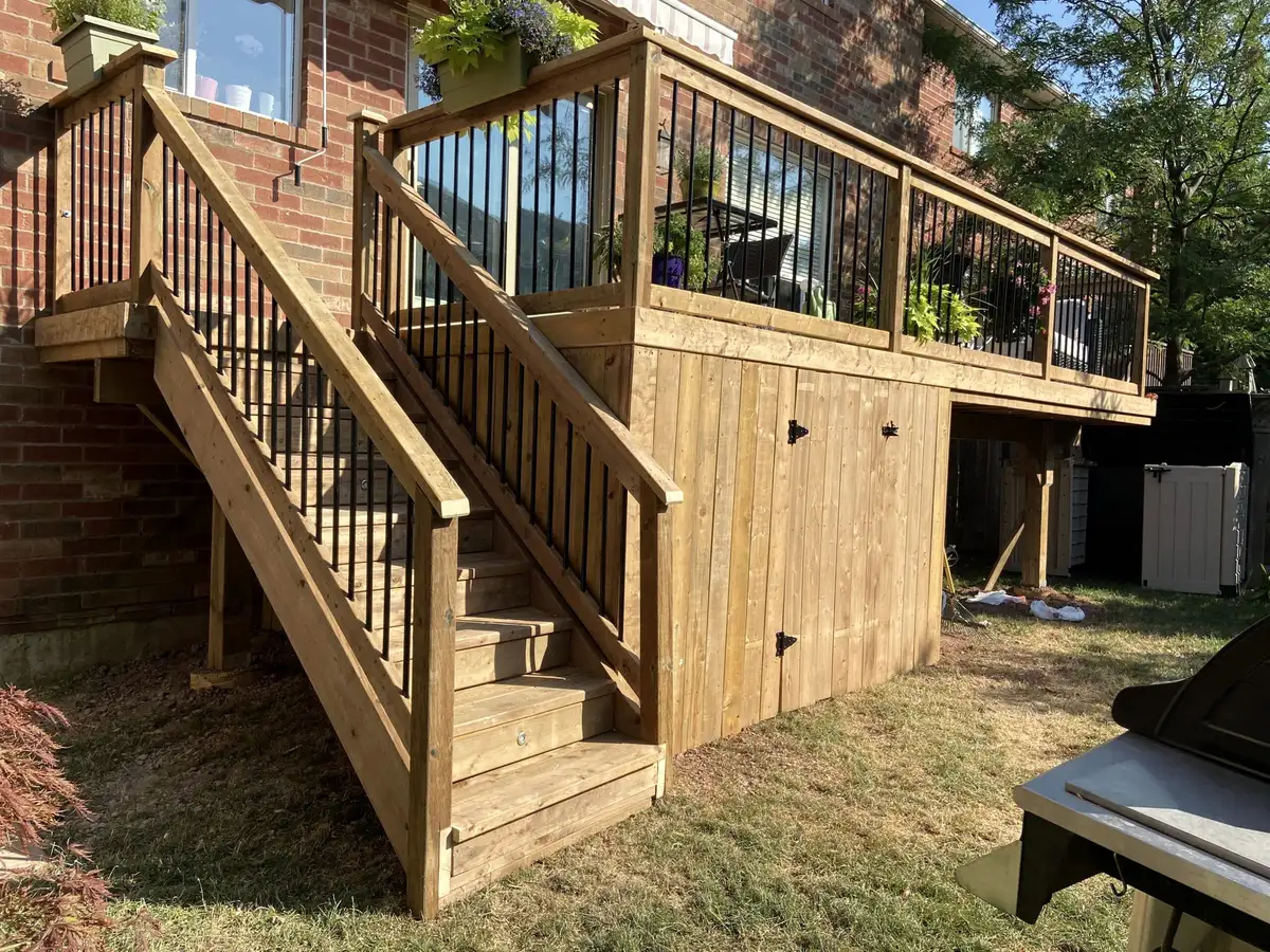 A pressure-treated stair turning at a landing up to a raised deck with black balusters, against a red-brick house.