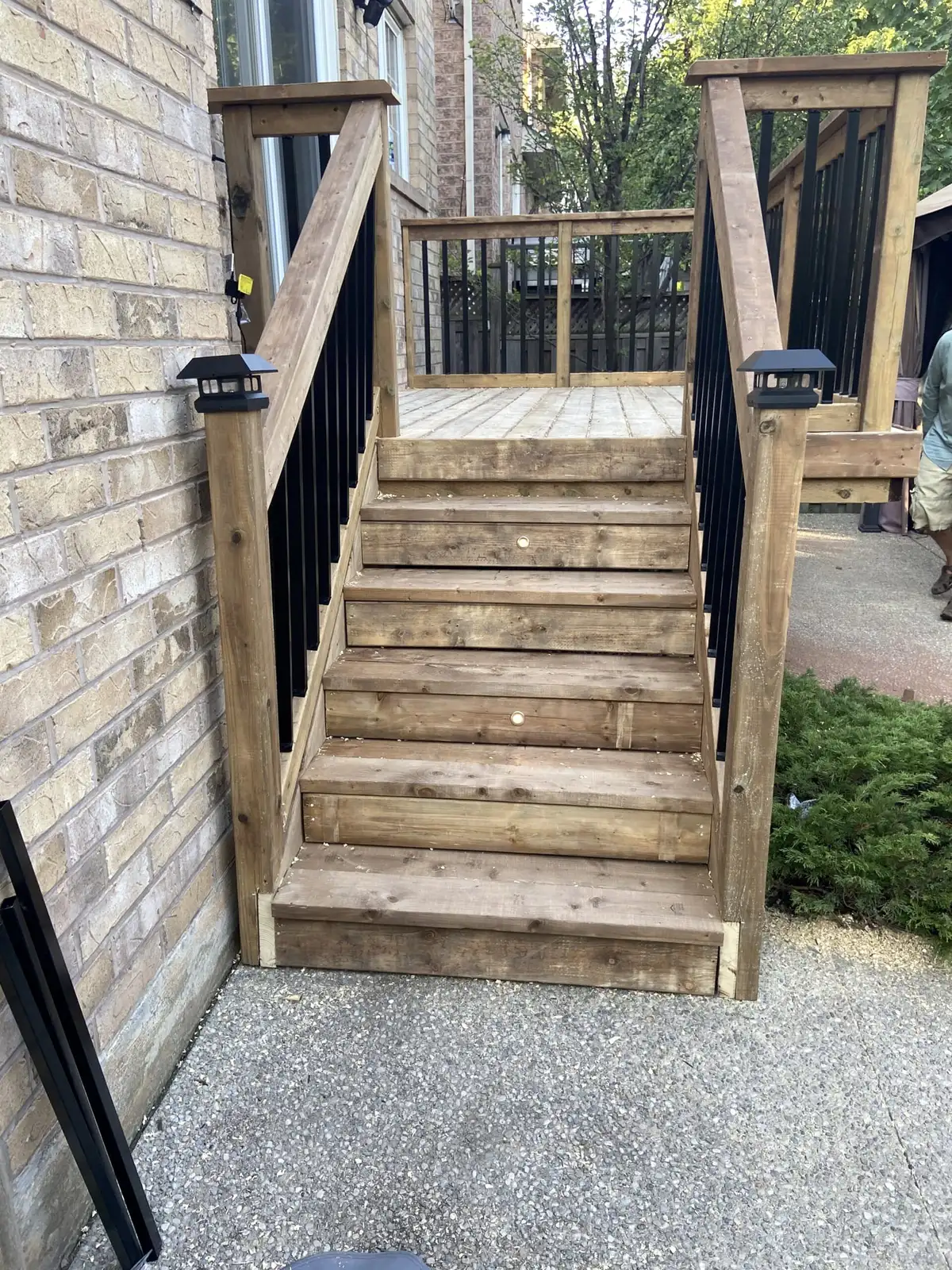 A straight pressure-treated wood stair with black balusters and solar caps on the posts, rising to an upper landing beside a buff-brick wall.